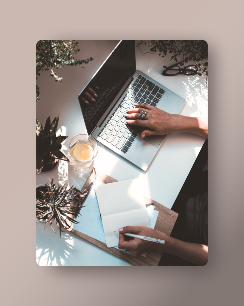 a woman in a modern office setting with lots of plants is typing on the computer with one hand and writing in a notebook in the other