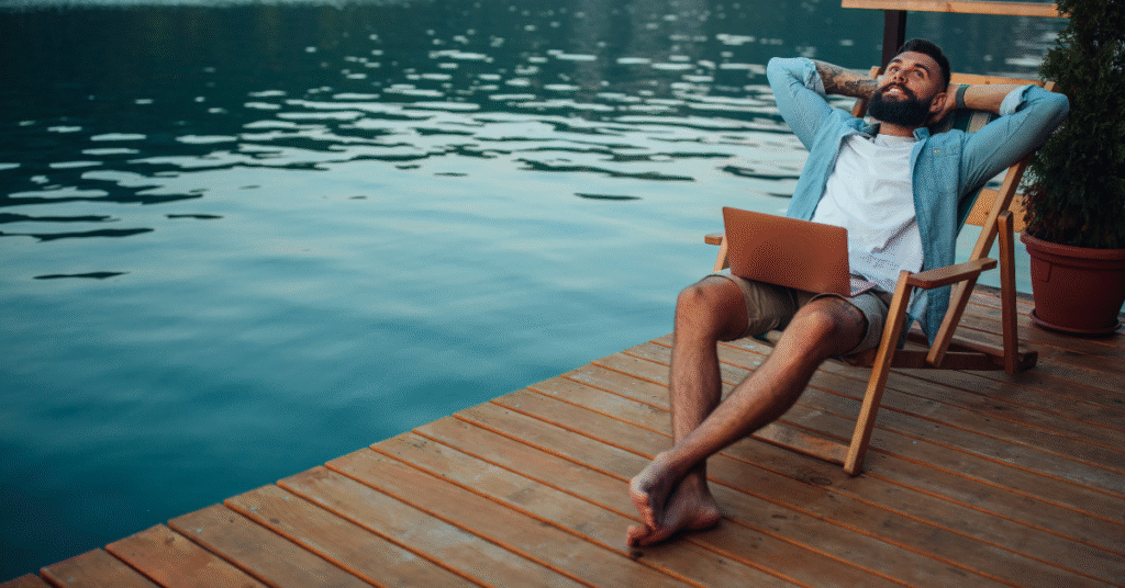 Man sitting on a wooden deck overlooking a lake, looking relaxed while working on his laptop in a peaceful outdoor setting