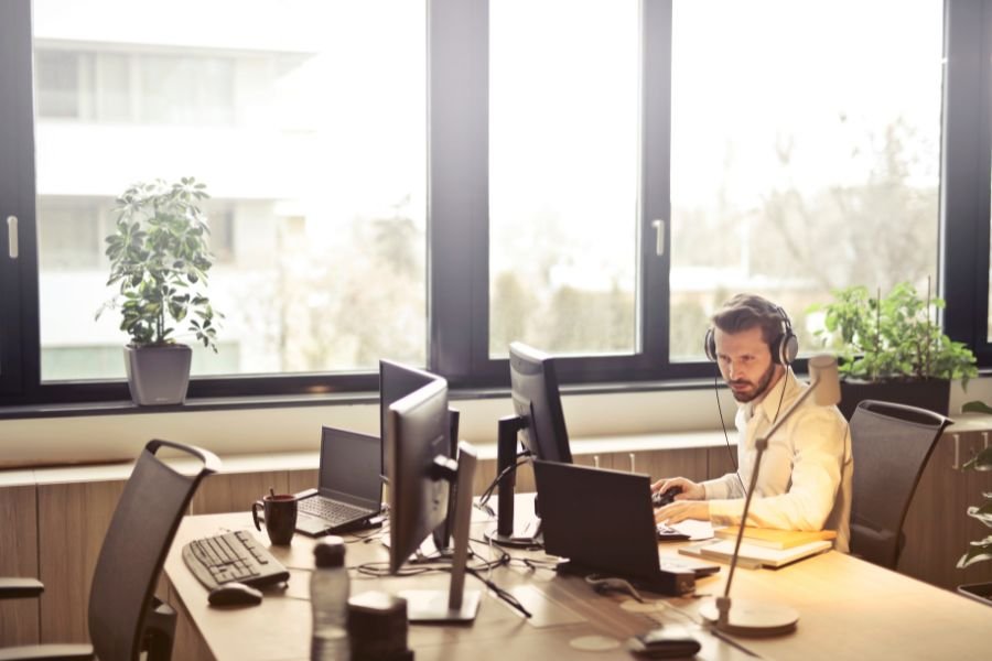 man sits in a large bright office space wearing headphones while looking at a computer screen