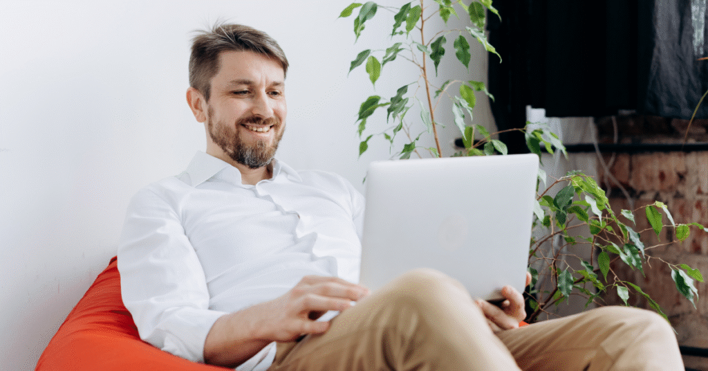 Smiling man sitting in an orange bean bag chair, working on a laptop in a relaxed, modern space