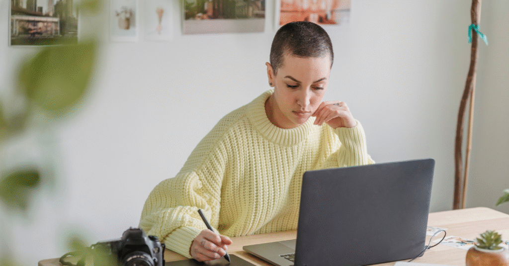 Woman with a shaved head wearing an oversized yellow sweater, working on a laptop in a bright, modern office space