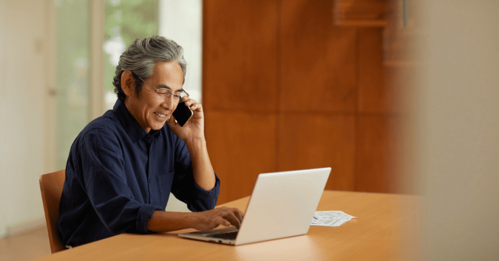 Person smiling while on a phone call and working on a laptop at a wooden desk, representing clear client communication and productive online collaboration.