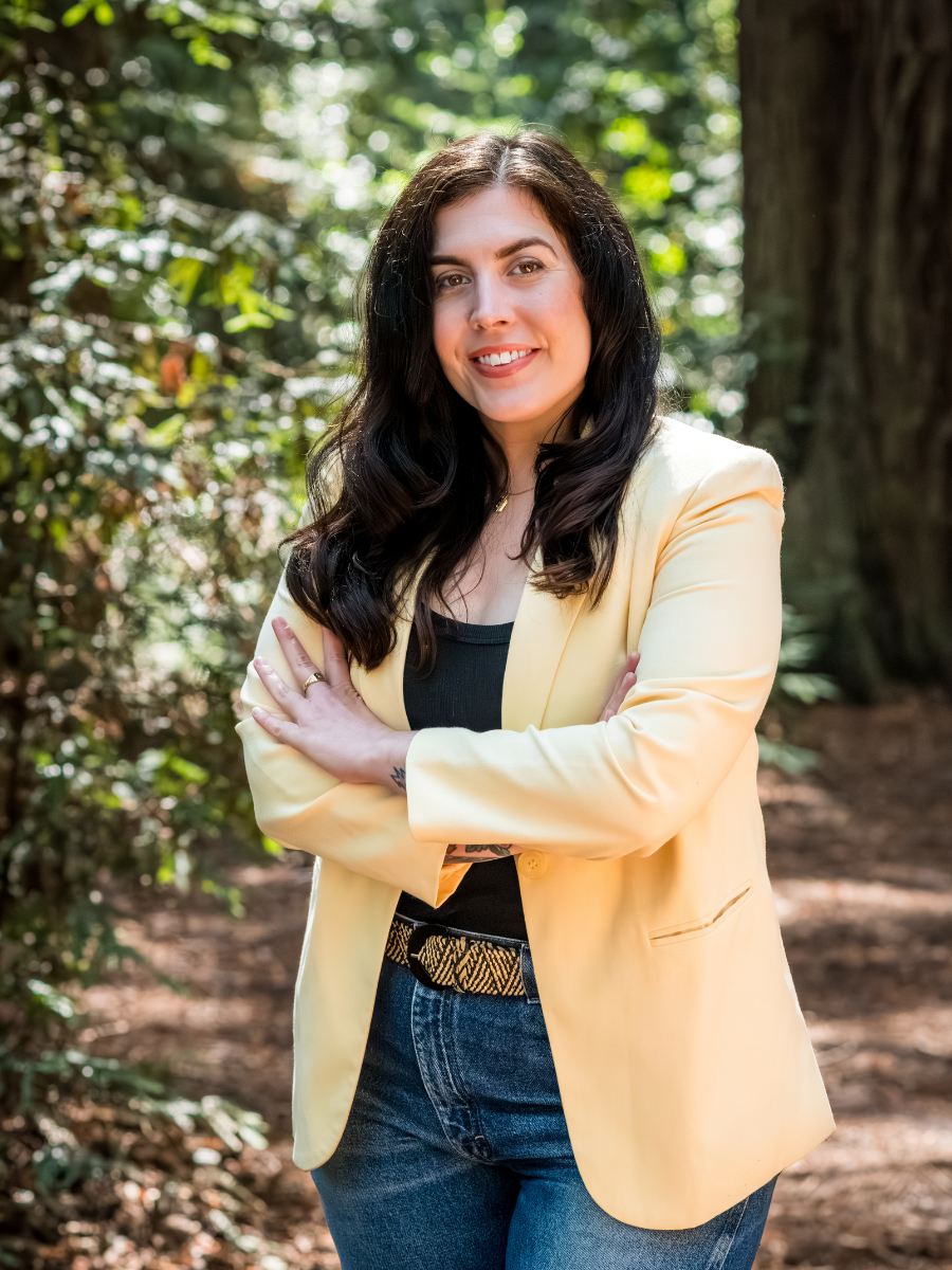 Lindsay Pfeiffer, founder of Bay Laurel Solutions, standing outdoors in a forest wearing a yellow blazer and jeans, smiling with arms crossed.