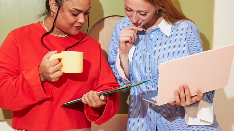 Two women reviewing brand materials and discussing design decisions while working on a laptop and clipboard.