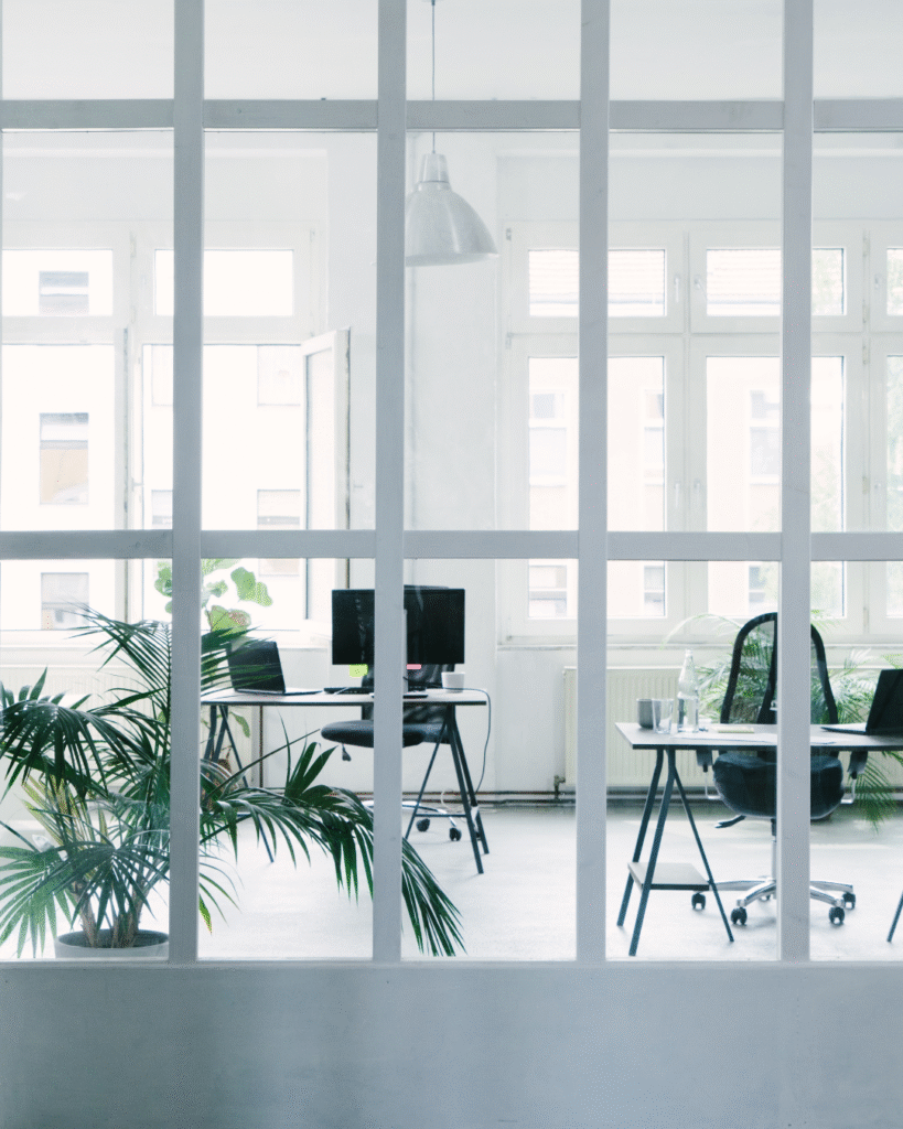 An empty, modern office seen through glass partitions, with desks, chairs, and plants in soft daylight.