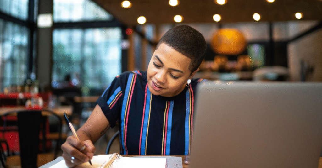 A woman sitting in a cafe writing her audience research checklist notes in a notebook with a laptop open beside her, representing the process of turning research insights into action.