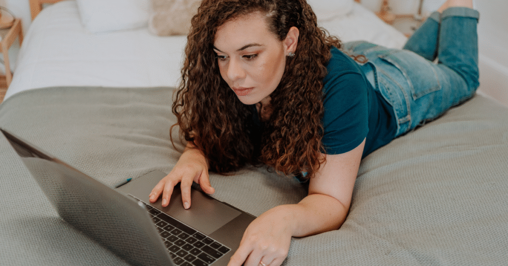 lady wearing jeans, laying down, scrolling on a laptop